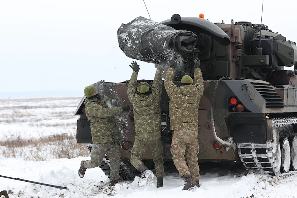 Three soldiers throw a large tarp in the back of a military vehicle moving through snow.